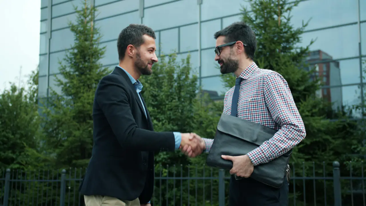 Two professionals shaking hands in front of an office building, one holding a portfolio